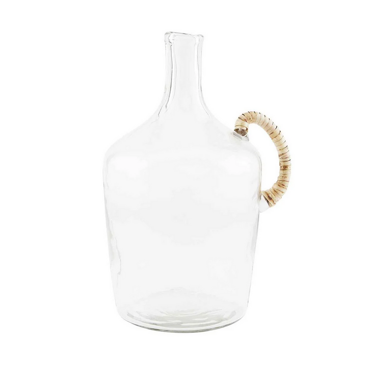A clear glass jug with a wicker wrapped handle placed on a white surface, with a larger glass jug in the background containing some dried plants.