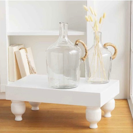 A clear glass jug with a wicker wrapped handle placed on a white surface, with a larger glass jug in the background containing some dried plants.