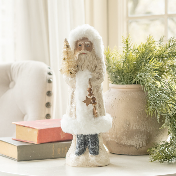 Decorative Santa figure of a bearded man in a white robe with stars, placed on a surface with books and a plant in the background.