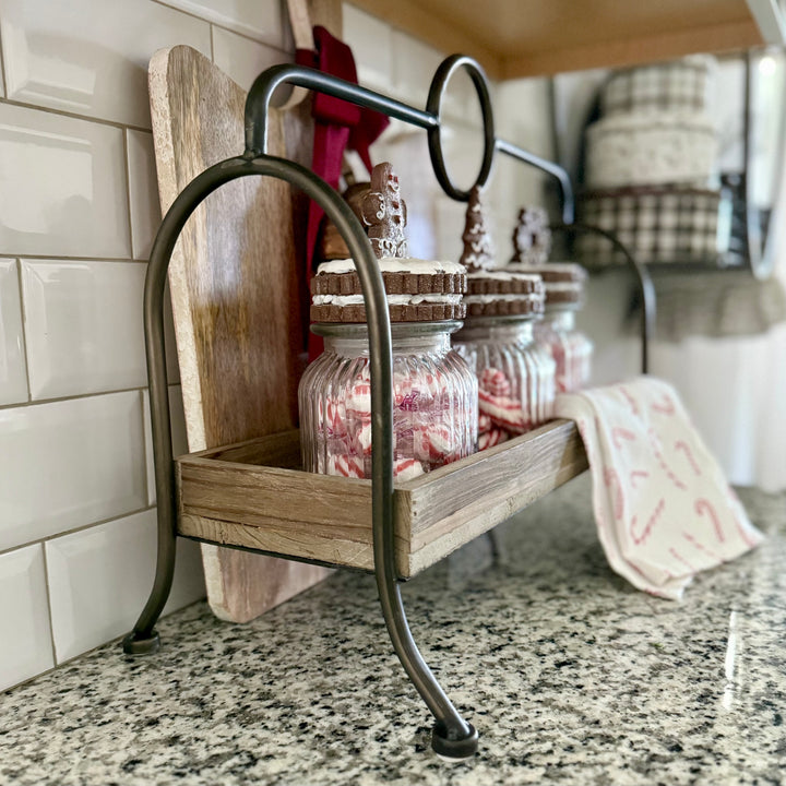 Wooden shelf with metal bracket holding glass jars with glittered gingerbread topper on a kitchen counter.