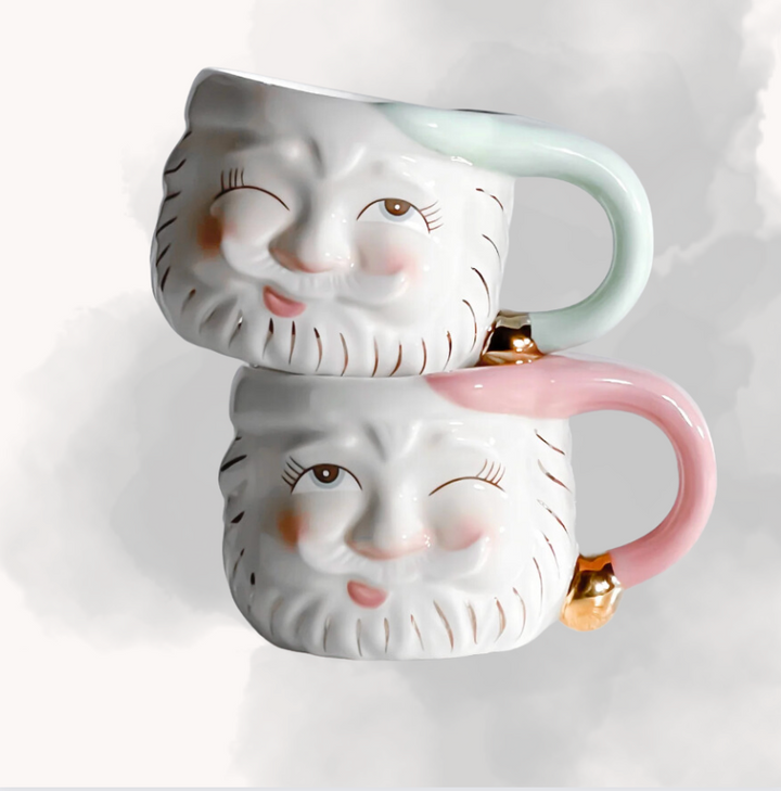 Two ceramic mugs with a whimsical Santa design, one in vintage mint color and the other in pink, both with gold accents.