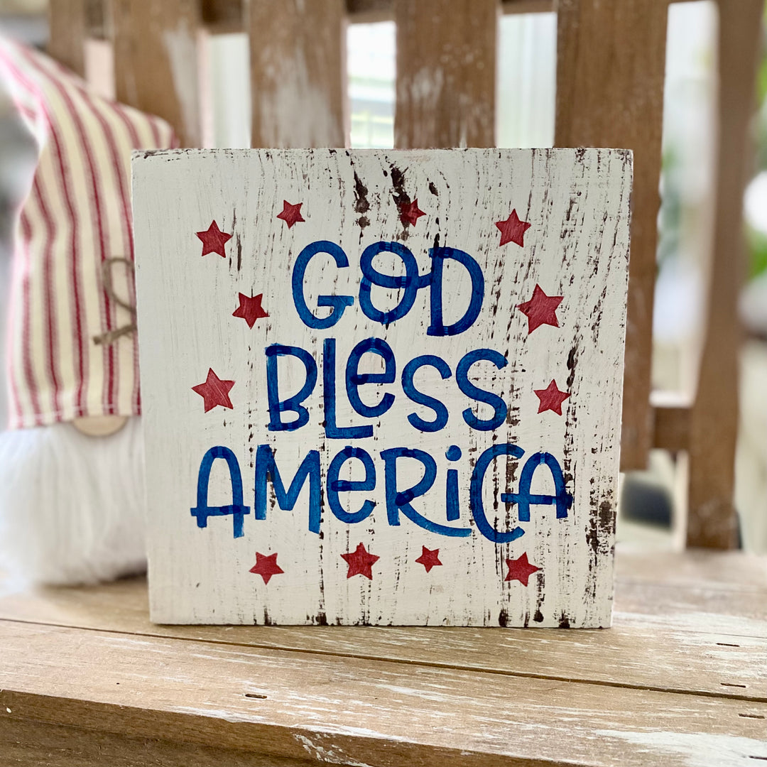 A decorative Americana themed arrangement featuring a wooden star, a gnome with a blue star hat, a small American flag, and a 'God Bless America' sign, all placed on a tiered tray.