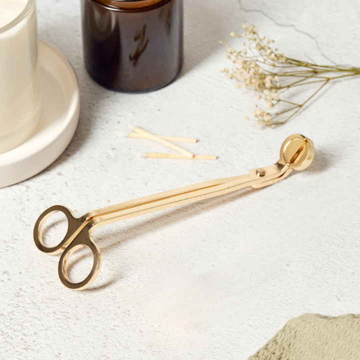 Gold metal candle wick trimmer with a circular handle, styled on a textured white surface with a white candle, amber glass jar, matchsticks, and dried flowers.