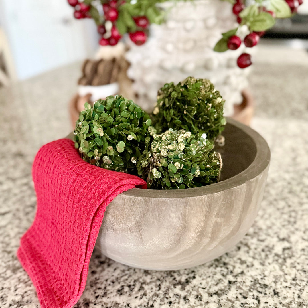 Dough bowl with glittered faux greenery and a red kitchen towel  on a countertop