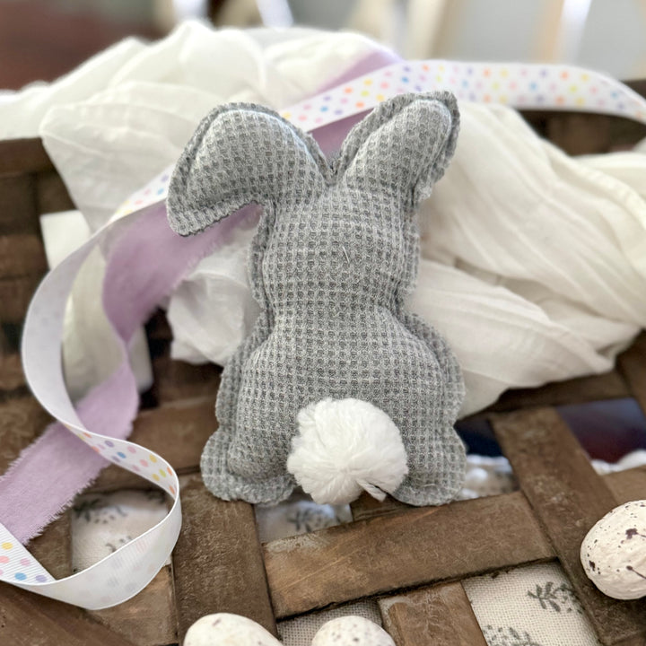 Gray textured bunny toy with white paws on a wooden surface with fabric and eggs.