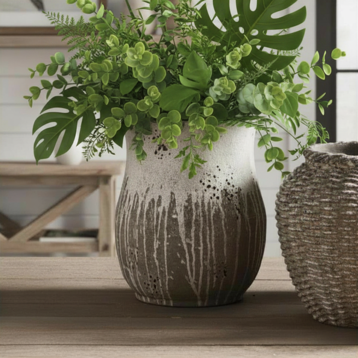 A handcrafted gray and white cement vase with a raw clay texture and thick glaze, displayed on a wooden surface with greenery.