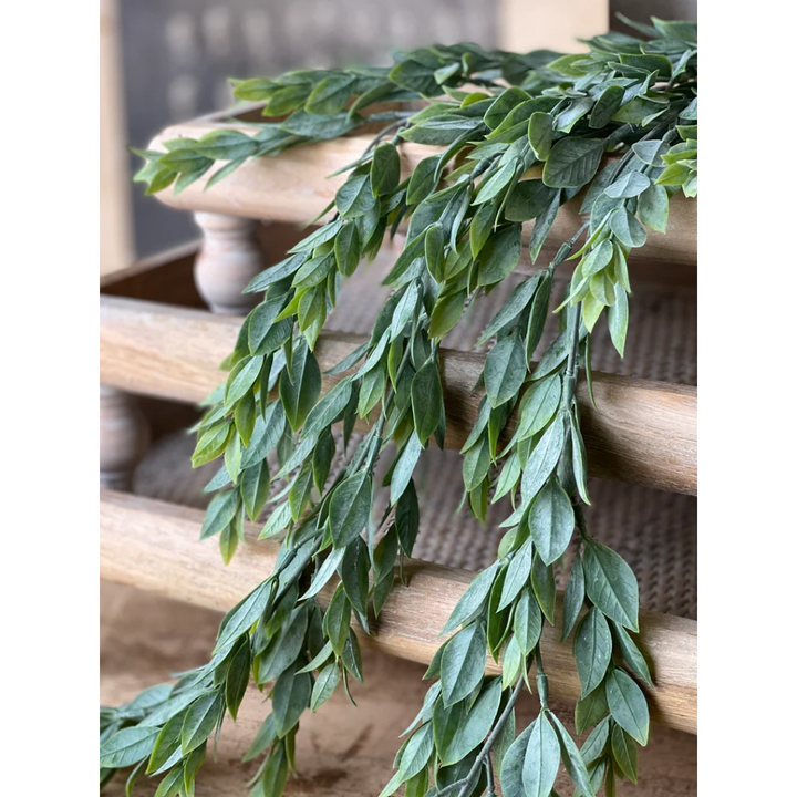 Green leafy vine branches draped over a light brown wooden railing or planter, photographed in a real setting with a blurred background.