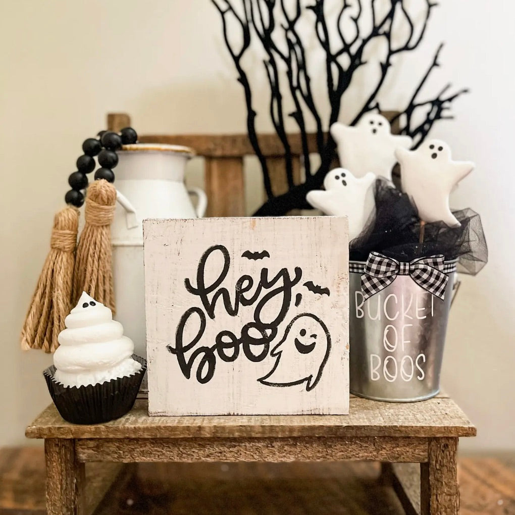 A rustic wood block sign with the phrase 'HEY BOO' painted in black, accompanied by decorative items such as a cupcakes, ghost-shaped cookies, and a bowl, all arranged on a wooden surface with a blurred background.