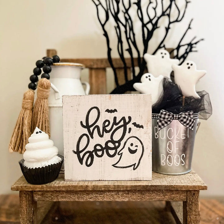 A rustic wood block sign with the phrase 'HEY BOO' painted in black, accompanied by decorative items such as a cupcakes, ghost-shaped cookies, and a bowl, all arranged on a wooden surface with a blurred background.