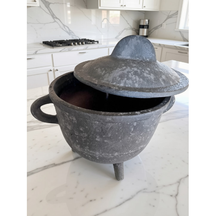 A dark gray cast aluminum lidded pot with a handle and tripod feet, sitting on a white marble kitchen countertop.