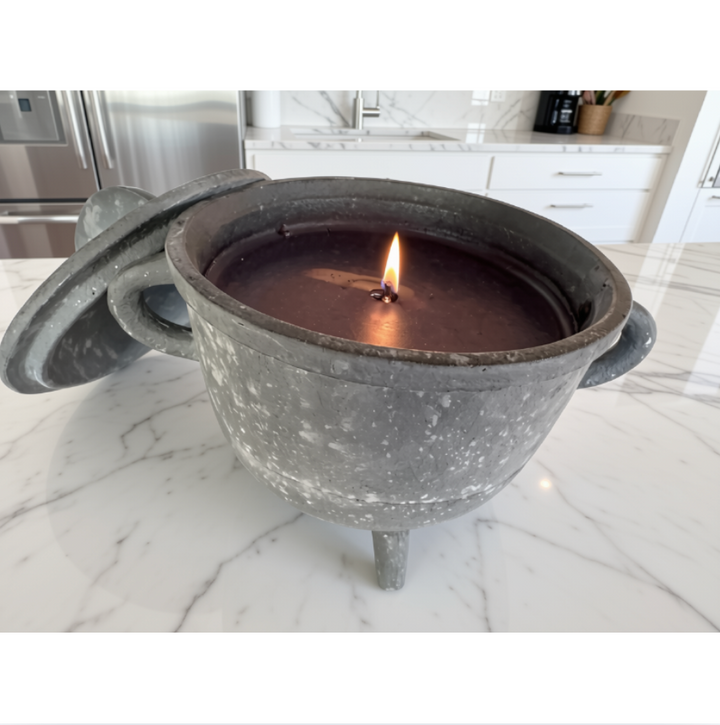A dark gray cast-aluminum style cauldron pot candle with a lid leaning against it, shown lit on a white marble kitchen countertop.