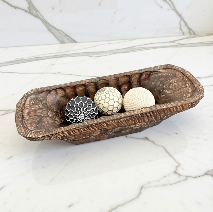 A rustic carved wooden bowl or tray holding three decorative textured balls, photographed on a white marble surface with a matching marble wall background.