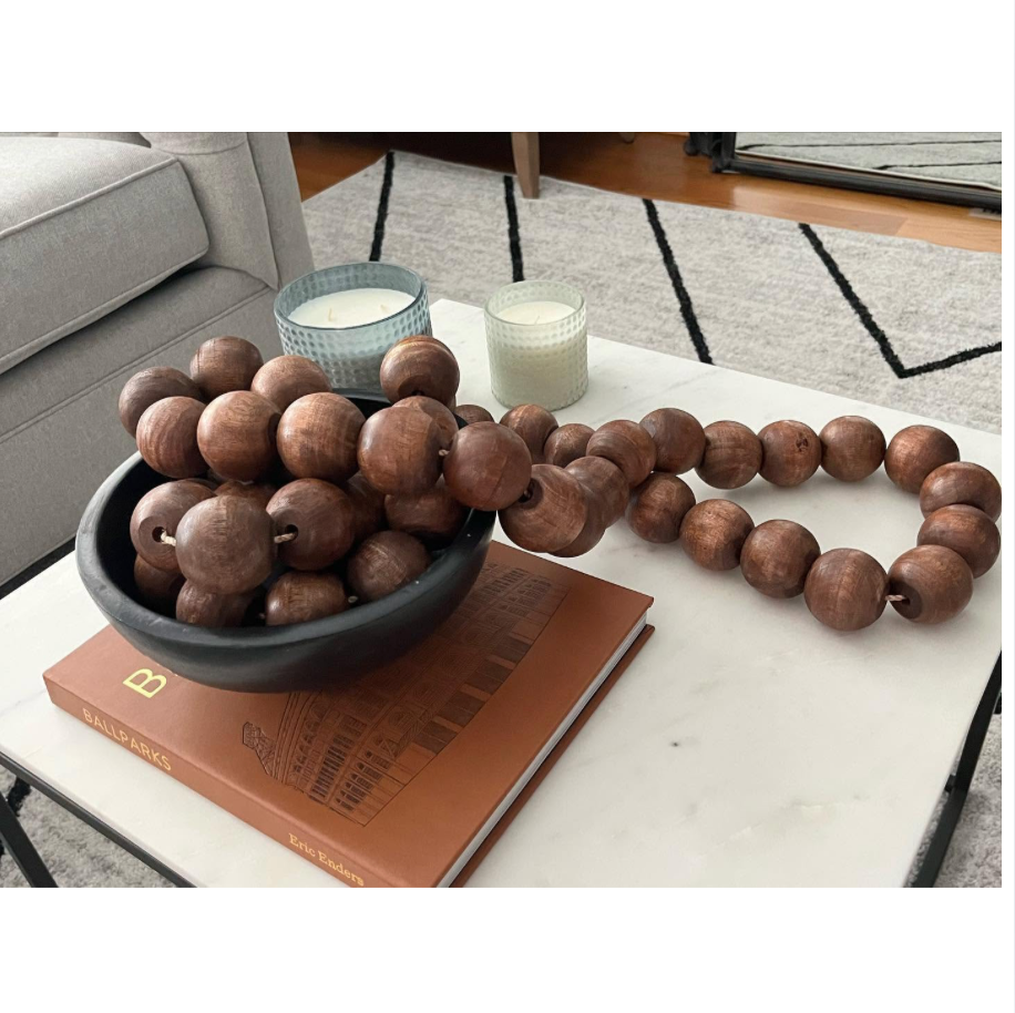 A long strand of medium brown wooden beads, some displayed in a black bowl and others laid out on a white marble coffee table, styled with a book, candles, and a sofa in a living room setting.