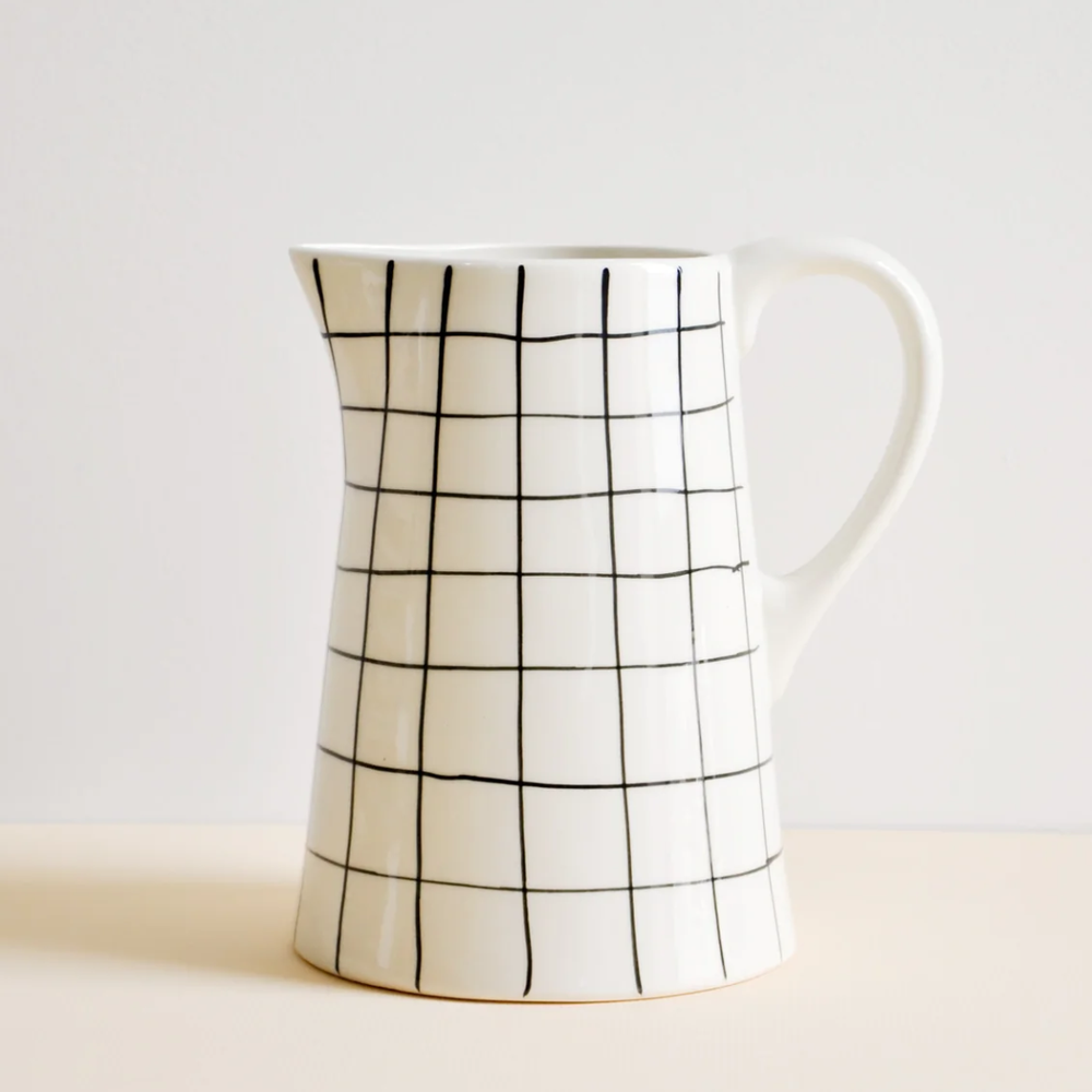 A white stoneware pitcher with a black hand-drawn grid pattern, standing on a light beige surface against a neutral off-white background.