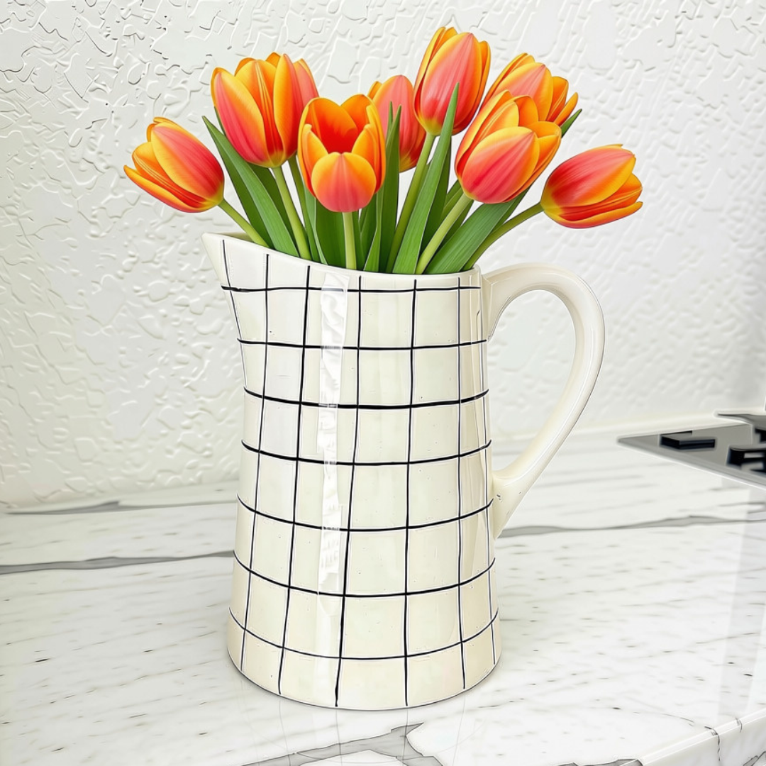 A white ceramic pitcher with a black grid pattern, filled with orange and pink tulips, set on a white marble countertop against a textured white wall.