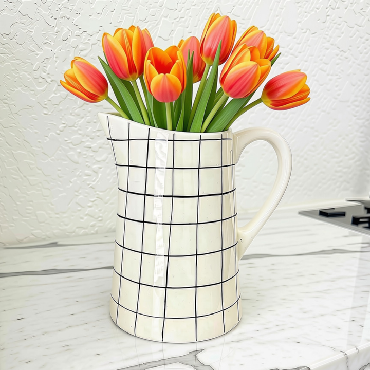 A white ceramic pitcher with a black grid pattern, filled with orange and pink tulips, set on a white marble countertop against a textured white wall.
