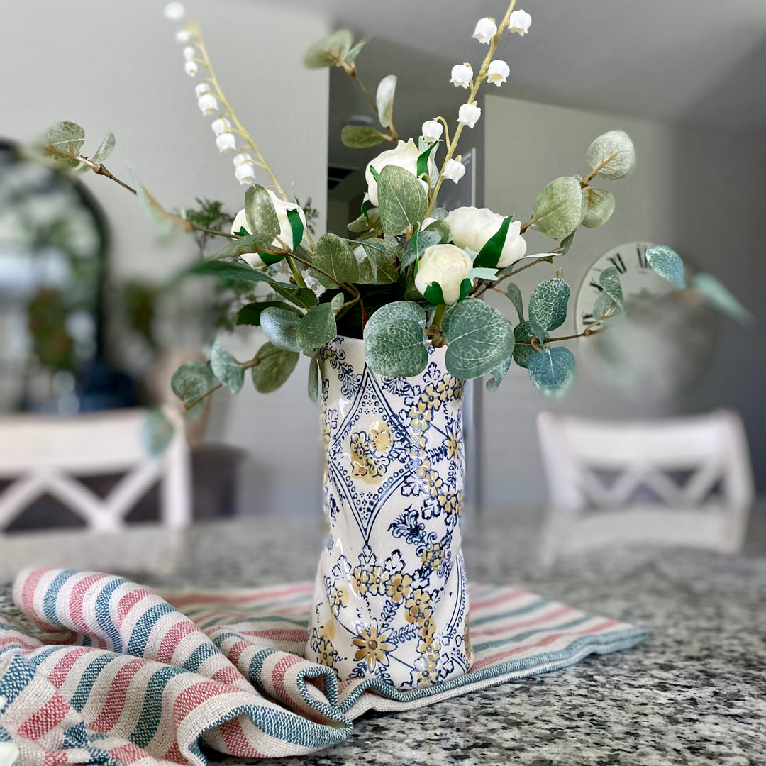 A hand-painted stoneware vase with a floral pattern, containing artificial greenery, placed on a table with a striped cloth in the foreground.