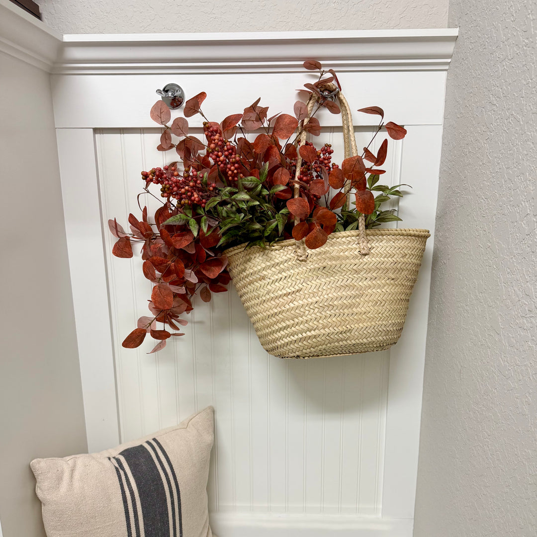 Decorative basket with red and green foliage attached to a white wall, with a pillow on a surface below.