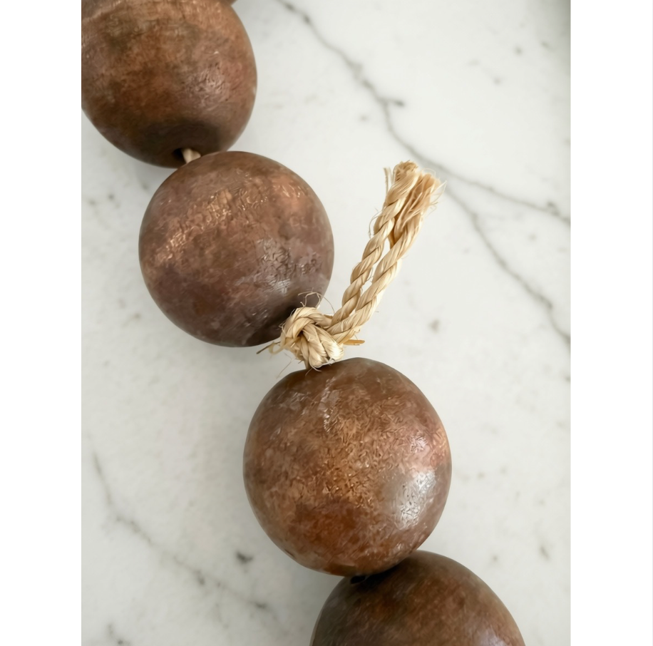 A close-up of a rustic beaded necklace or garland made of large, round, textured brown wooden beads strung on a natural tan twine cord, set against a white marble background.