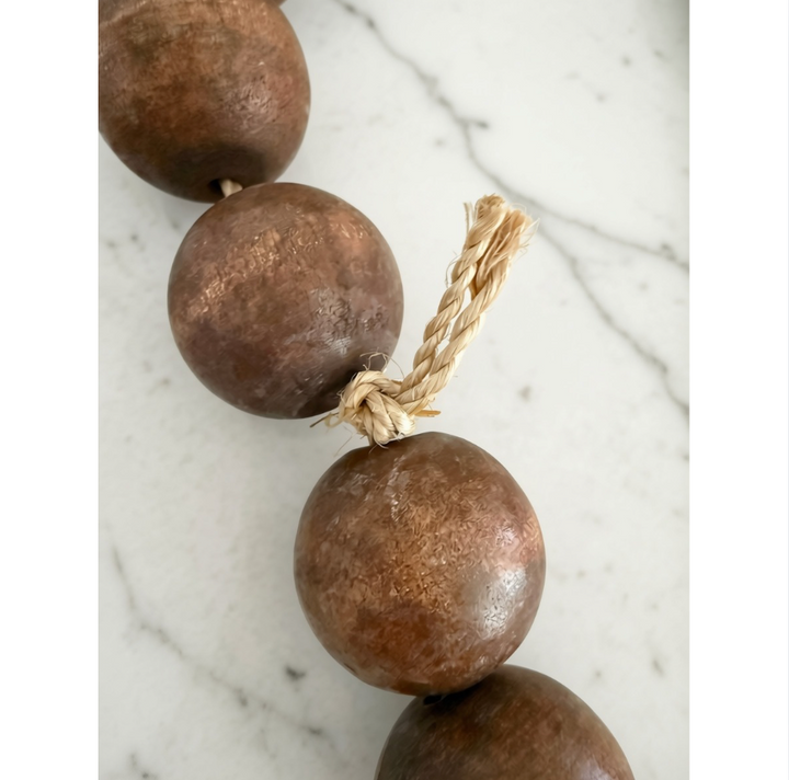 A close-up of a rustic beaded necklace or garland made of large, round, textured brown wooden beads strung on a natural tan twine cord, set against a white marble background.