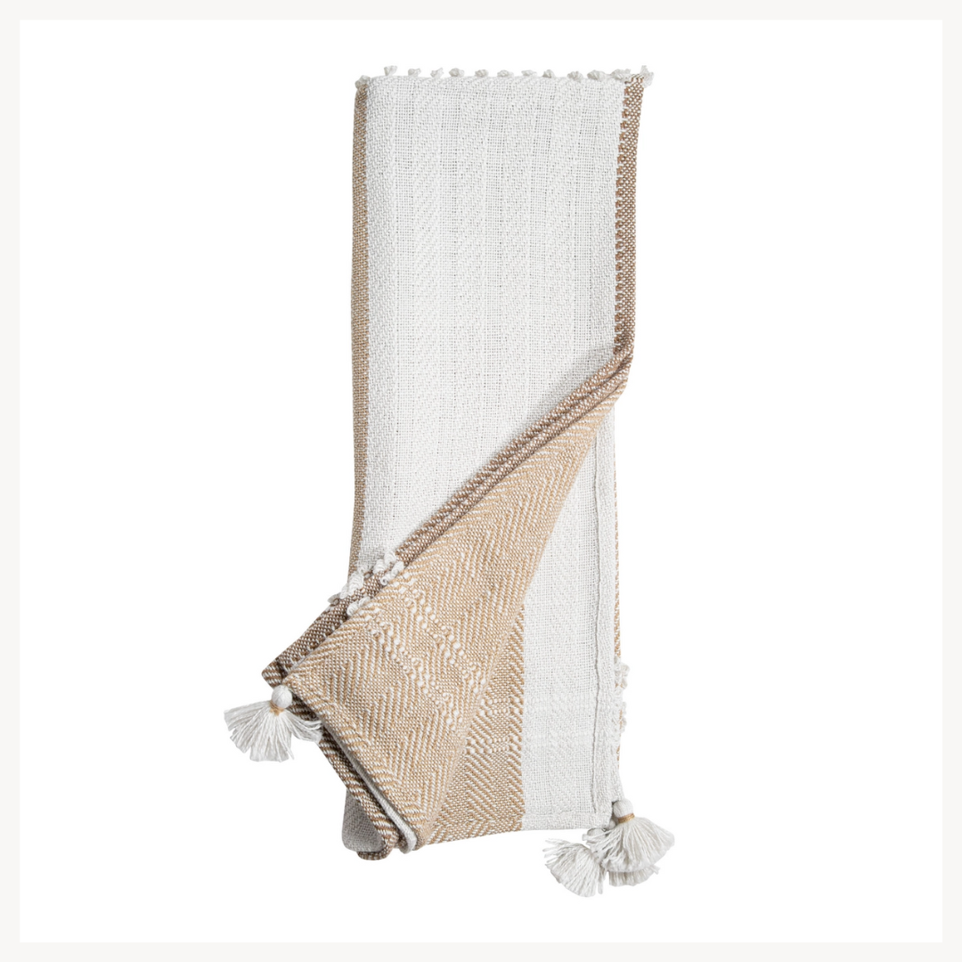 A folded hand woven cotton throw blanket with a patchwork plaid design in beige and brown colors, featuring corner tassels.