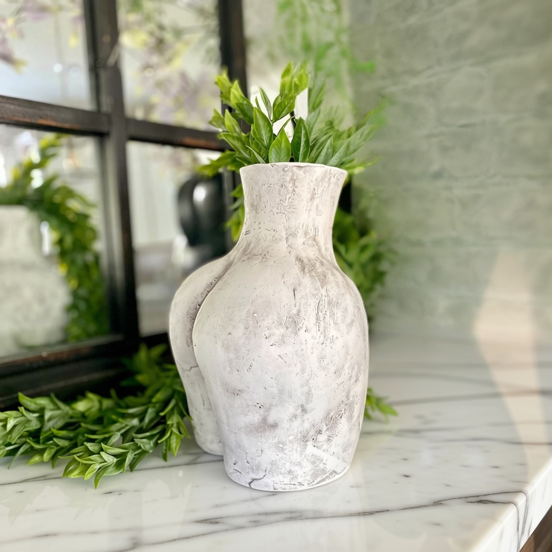 A textured, distressed white ceramic vase with a narrow neck and wide rounded body, filled with green foliage, displayed on a white marble surface.