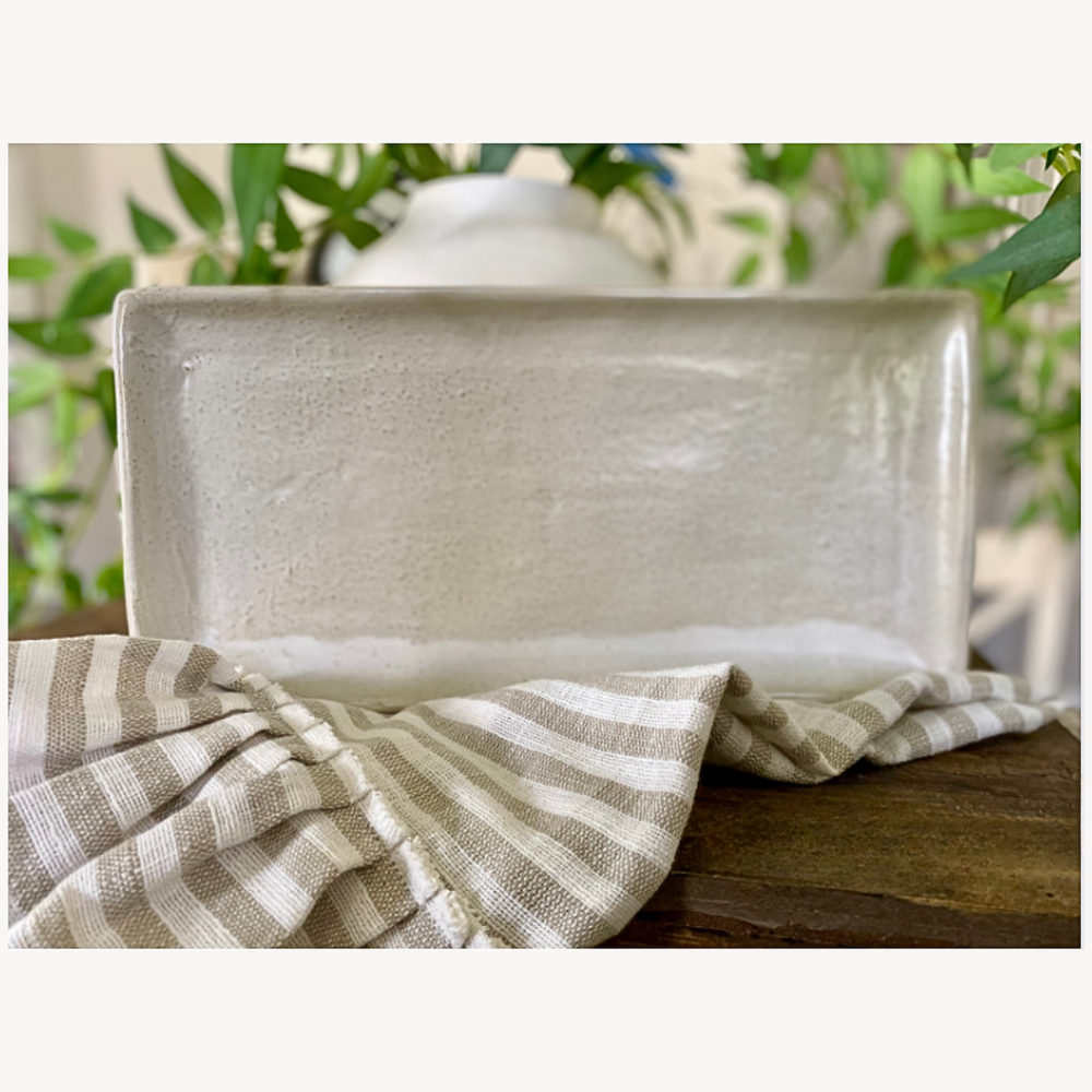 Rectangular stoneware tray with a striped tea towel on a wooden surface, blurred greenery in the background