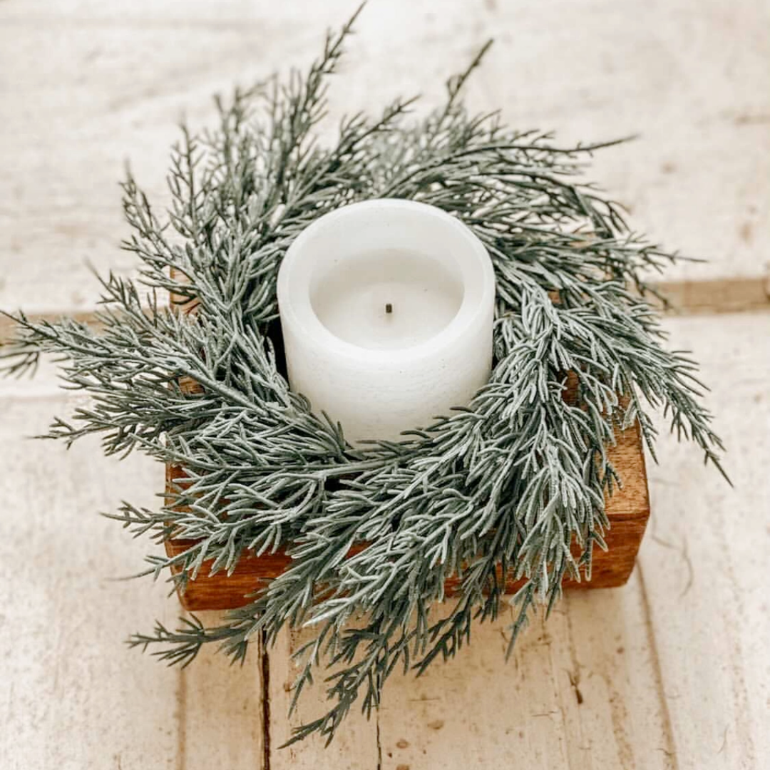 White candle in a wooden holder surrounded by greenery on a light wooden surface
