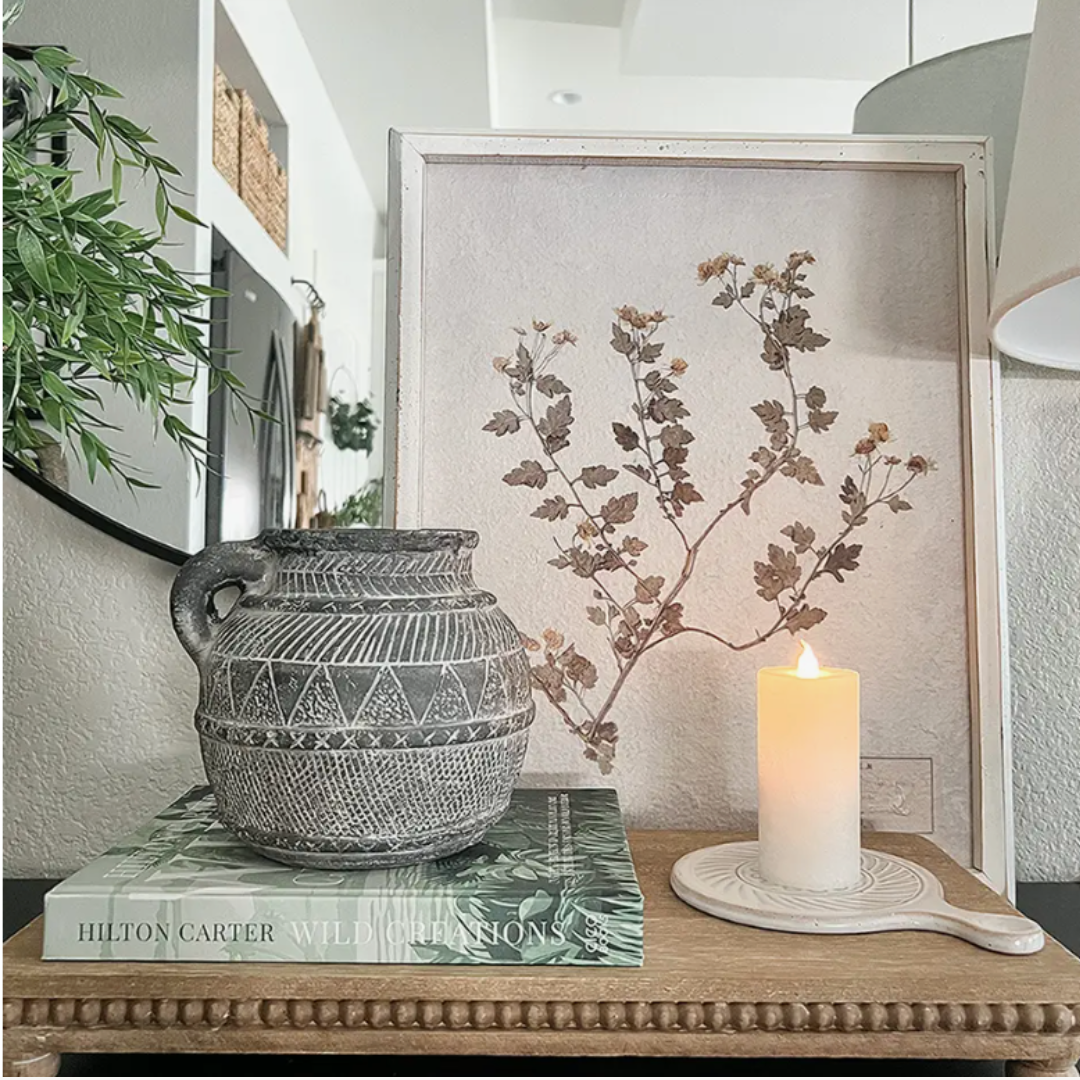 A handmade gray terracotta vase with an Aztec-inspired etched pattern, displayed on a wooden surface alongside a framed plant print and a candle.