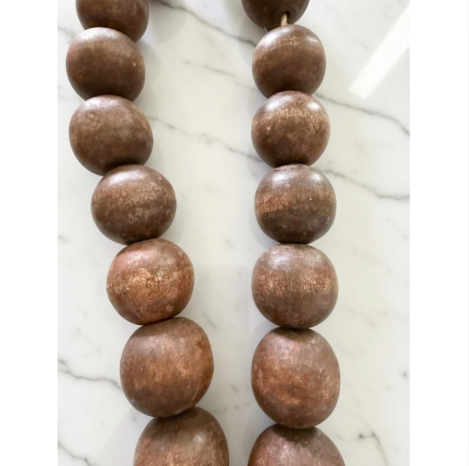 A close-up of a beaded necklace or strand of large round wooden beads in a warm medium brown tone, laid on a white marble surface.