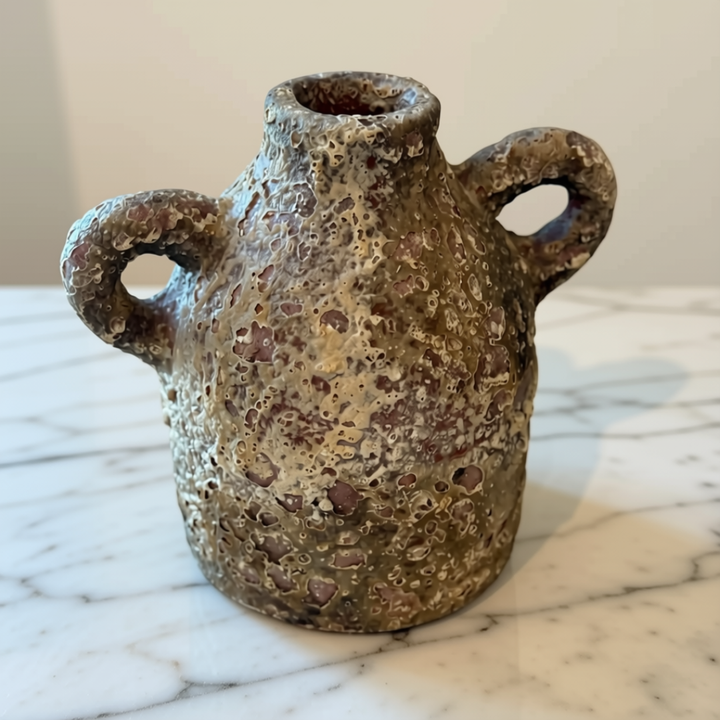 A small handmade stone vase with a textured, porous surface and two loop handles, sitting on a white marble surface against a neutral beige background.