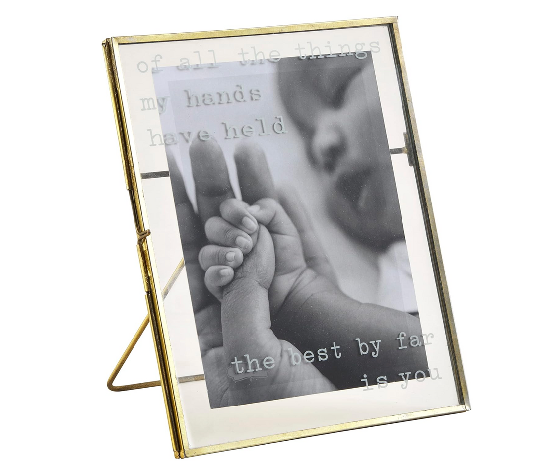 A brass finished metal picture frame with pressed glass panes, displaying a photo and the text 'of all the hands have held the best by far is you'.