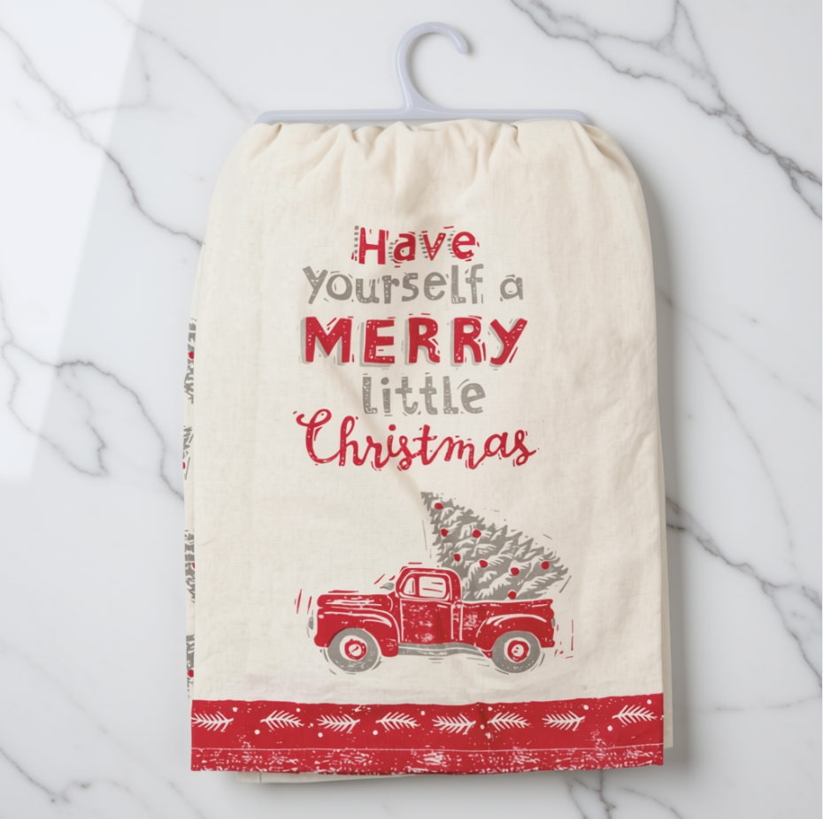A cream-colored cotton tea towel with a festive Christmas design, hanging on a white plastic hanger against a white marble background. The towel features the text 'Have yourself a MERRY little Christmas' in red and gray lettering, above an illustration of a red truck carrying a Christmas tree, and finishes with a red patterned border at the bottom.