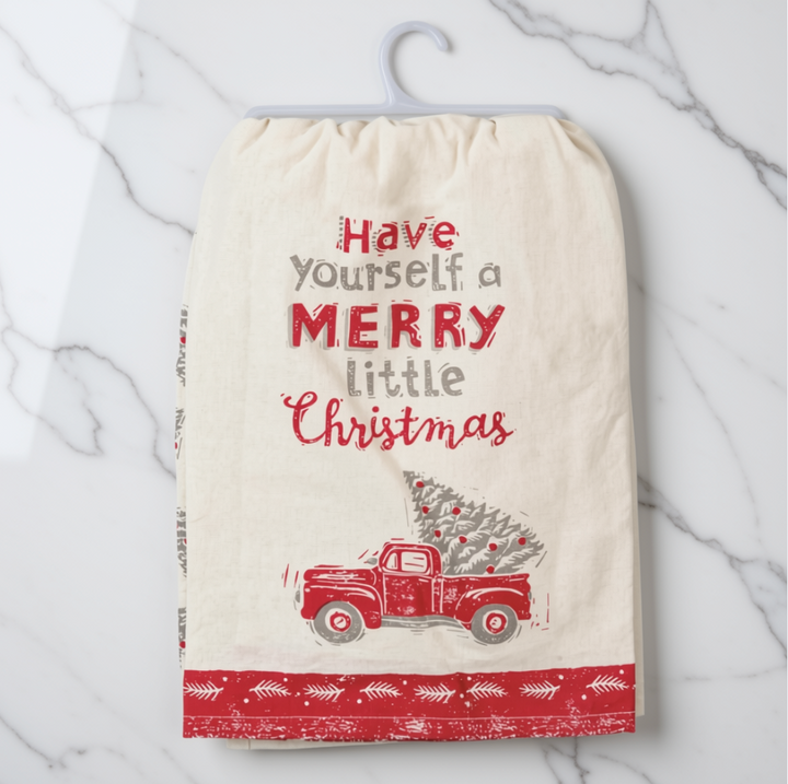 A cream-colored cotton tea towel with a festive Christmas design, hanging on a white plastic hanger against a white marble background. The towel features the text 'Have yourself a MERRY little Christmas' in red and gray lettering, above an illustration of a red truck carrying a Christmas tree, and finishes with a red patterned border at the bottom.