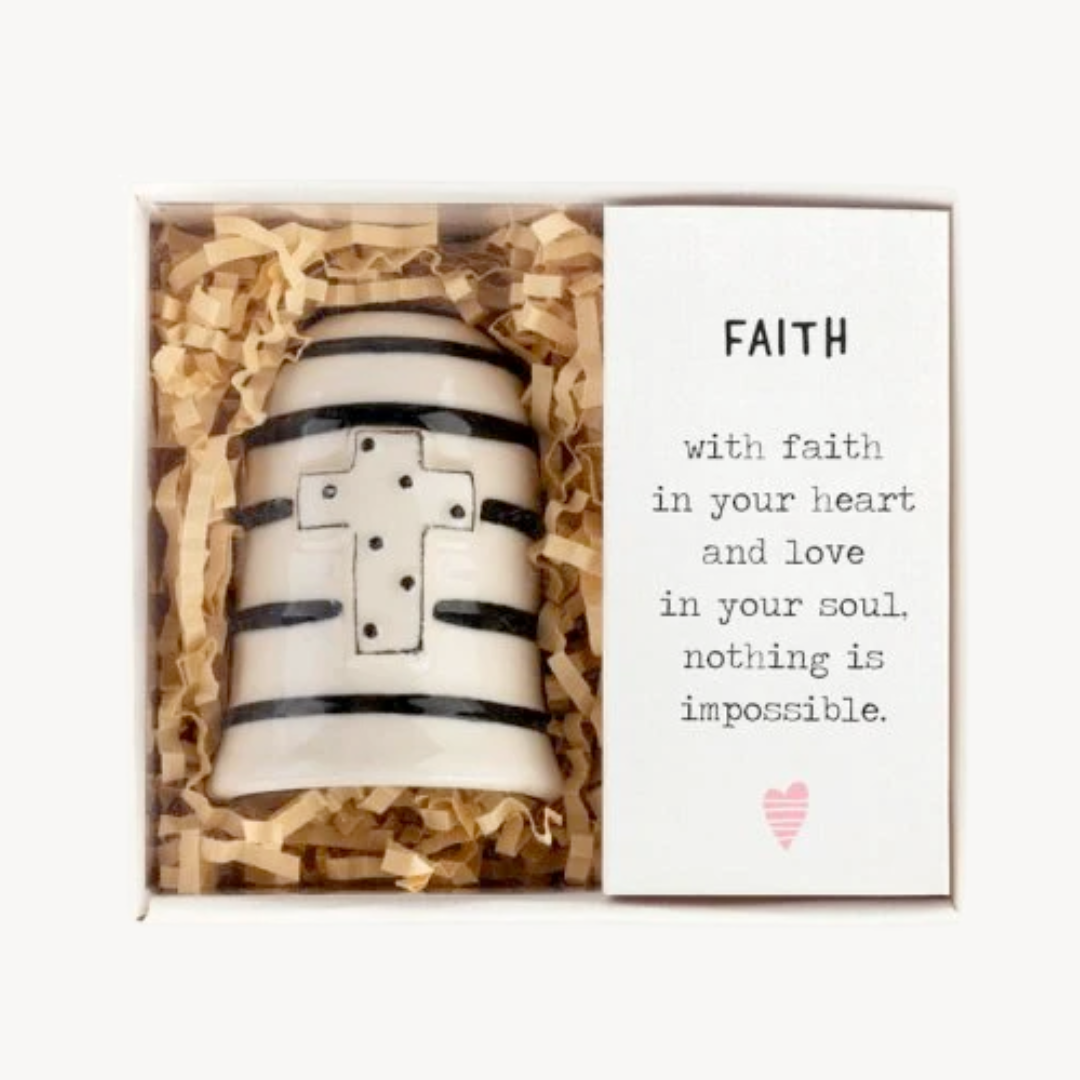 A hand-painted white stoneware bell with black stripes and a black and white cross, displayed in a box with wooden shavings, alongside a card with an inspirational message about faith.