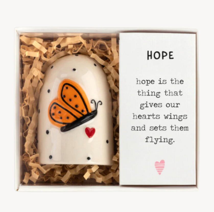 A white bell with black polka dots, an orange butterfly, and a small red heart underneath. The bell comes in a box with a 'HOPE' card and is surrounded by packing shavings.