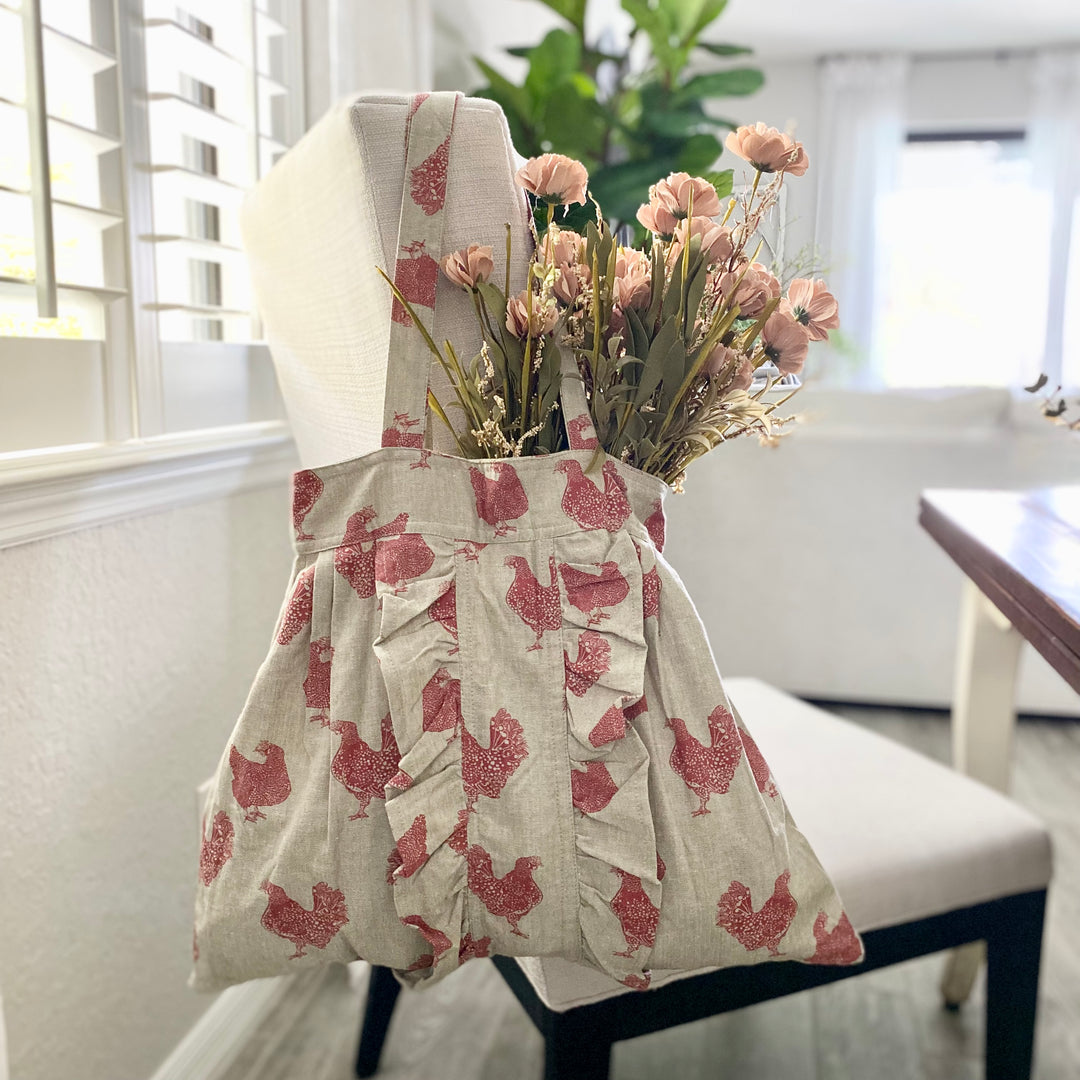 A beige shopping tote with a red chicken pattern, displayed in a home setting with a vase of dried flowers on top.