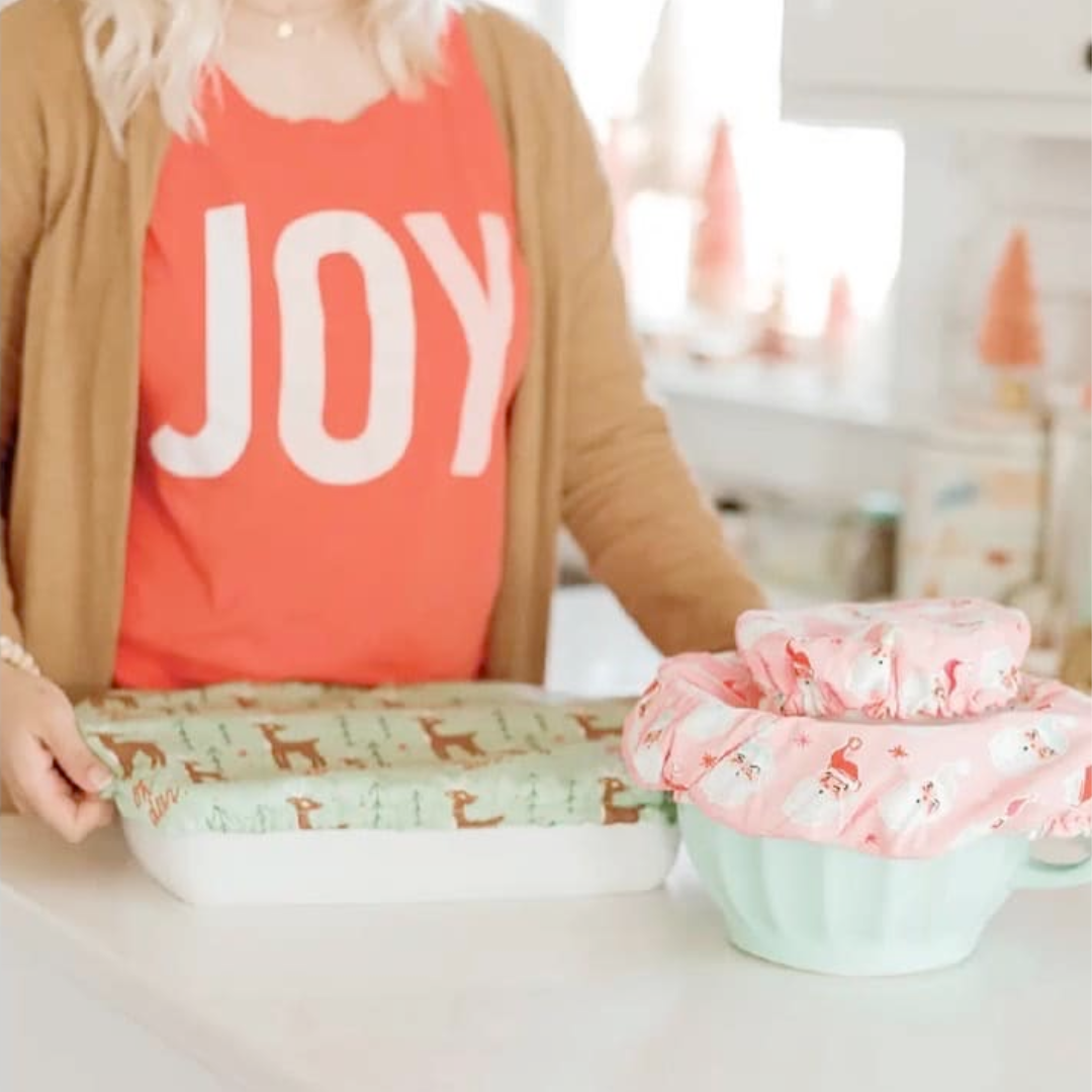 A person holding a green fabric bowl cover with a reindeer pattern. On the table are more bowl covers and a casserole dish.