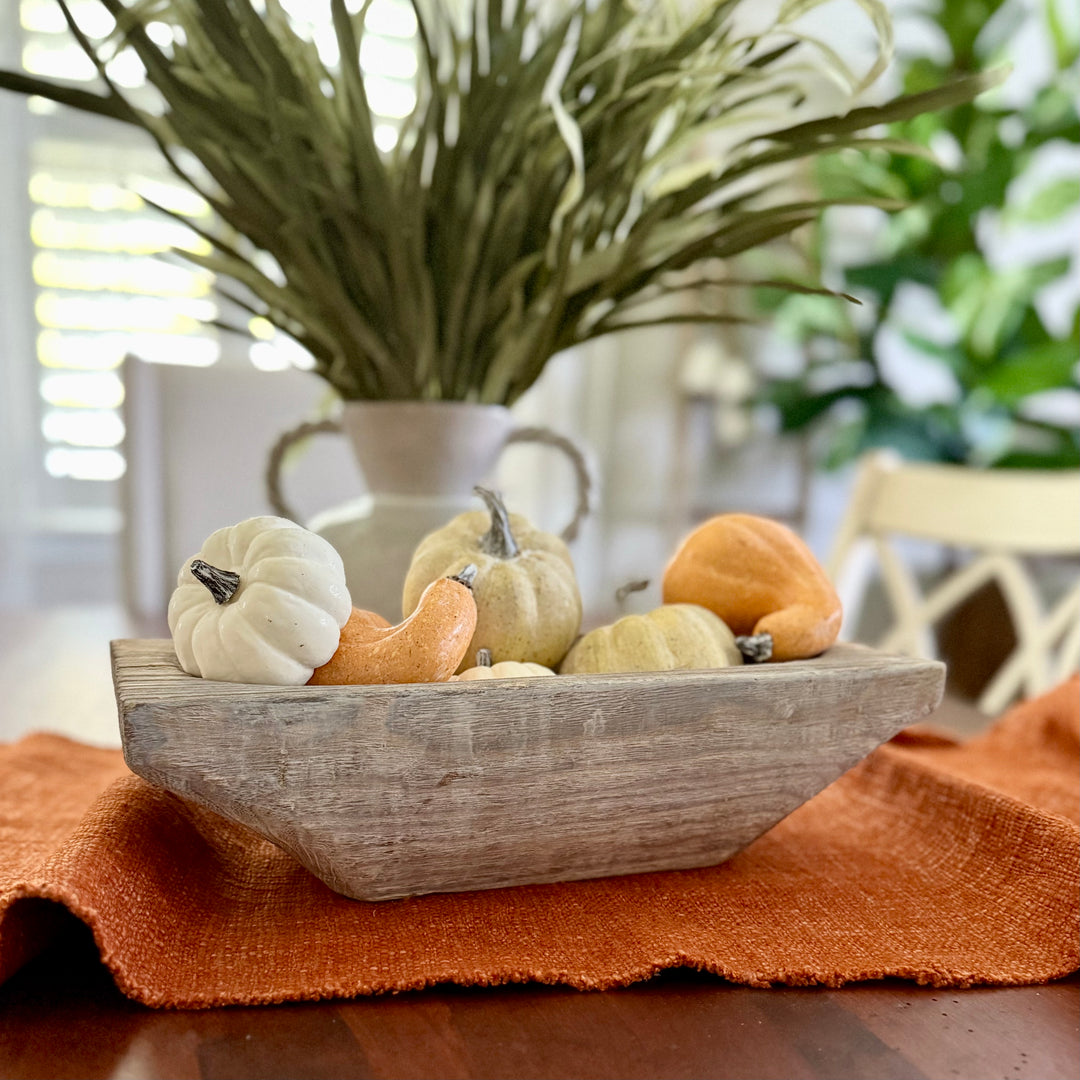 Decorative wooden bowl with small pumpkins and gourds on a table with a plant in the background.