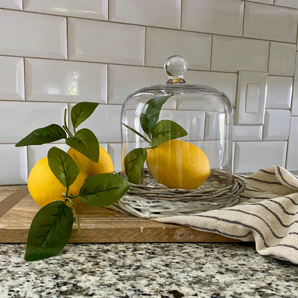 A set of three artificial lemons with attached green foliage, placed on a wooden tray, with one lemon under a clear glass cloche.