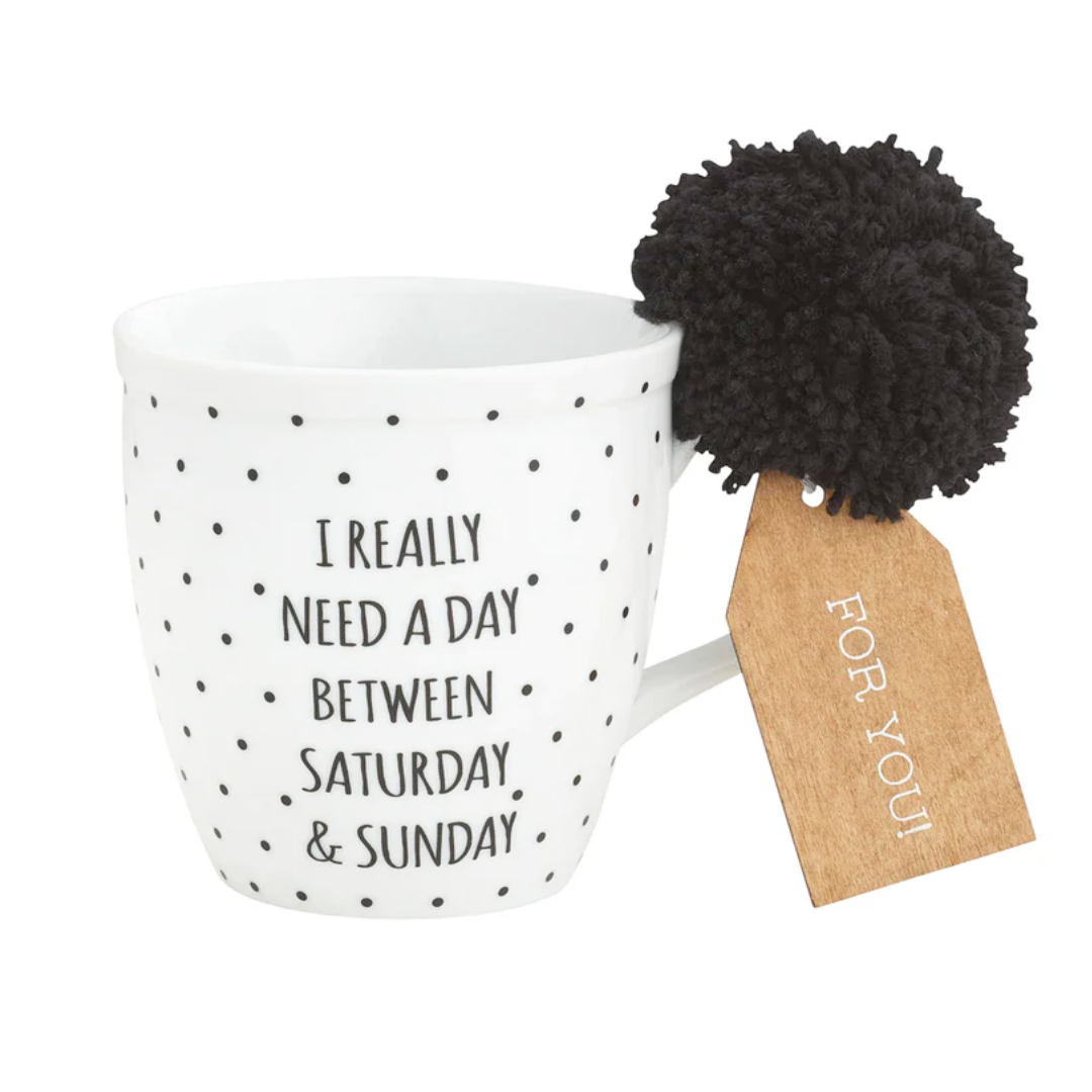 A white porcelain mug with black text and polka dots, featuring the phrase 'I Really Need A Day Between Saturday & Sunday', accompanied by a black pom pom and a wooden gift tag.