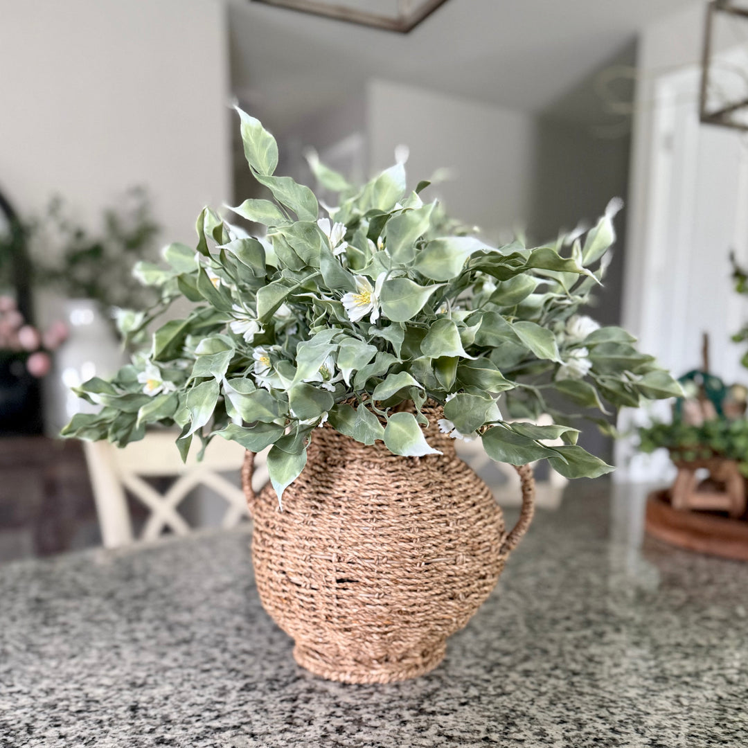 Artificial greenery stems in a woven vase on a table with a blurred indoor background
