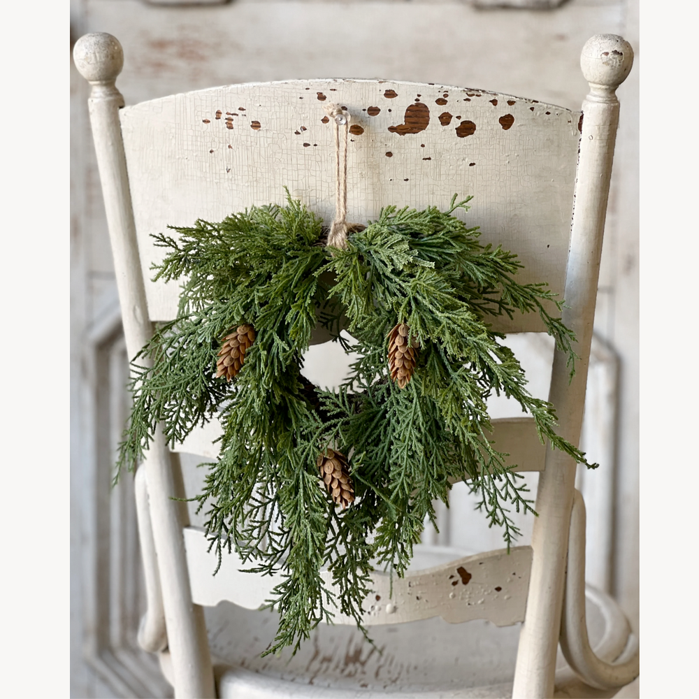 Mini green cedar wreath with pine cones hanging on a white wooden chair.