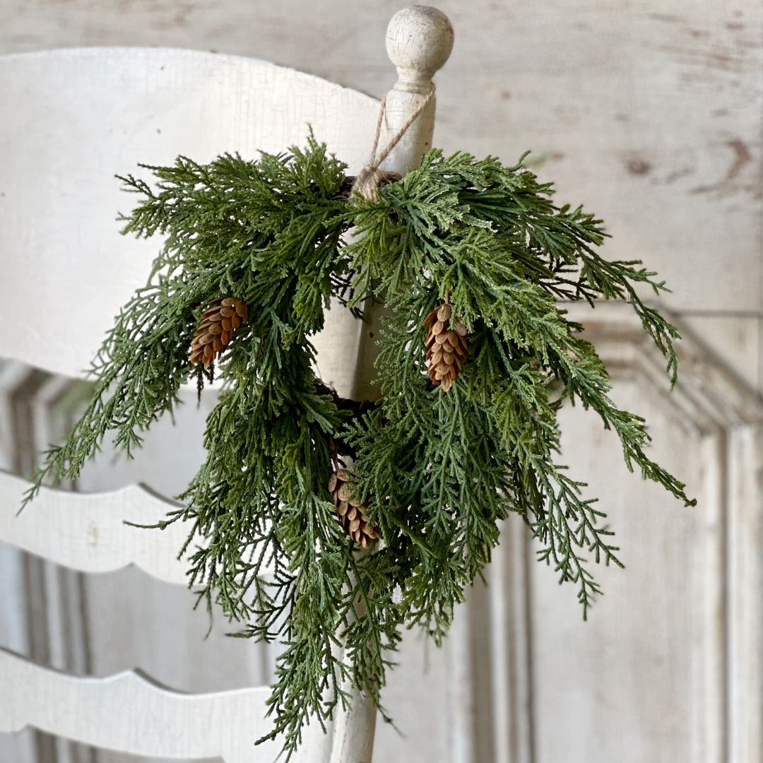 Green cedar  wreath with pine cones on a white chair against a wooden wall.