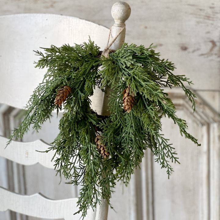 Green cedar  wreath with pine cones on a white chair against a wooden wall.