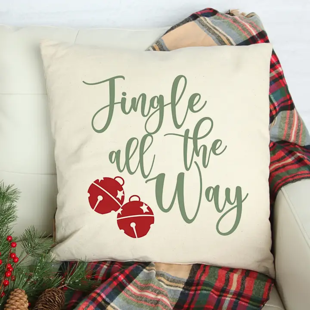 Decorative pillow with 'Jingle all the Way' text and jingle bells on a couch with plaid blanket and Christmas decor.