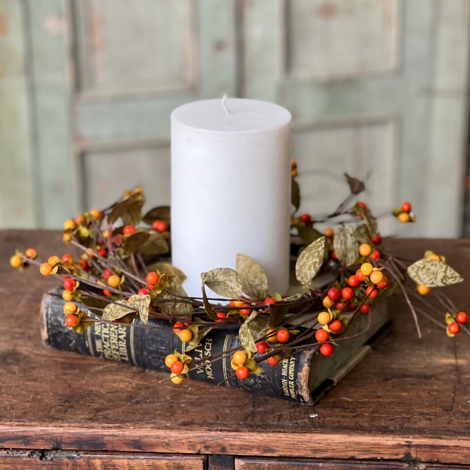 A candle ring with a greenery and bittersweet berry decoration, holding a white candle, on a wooden surface.