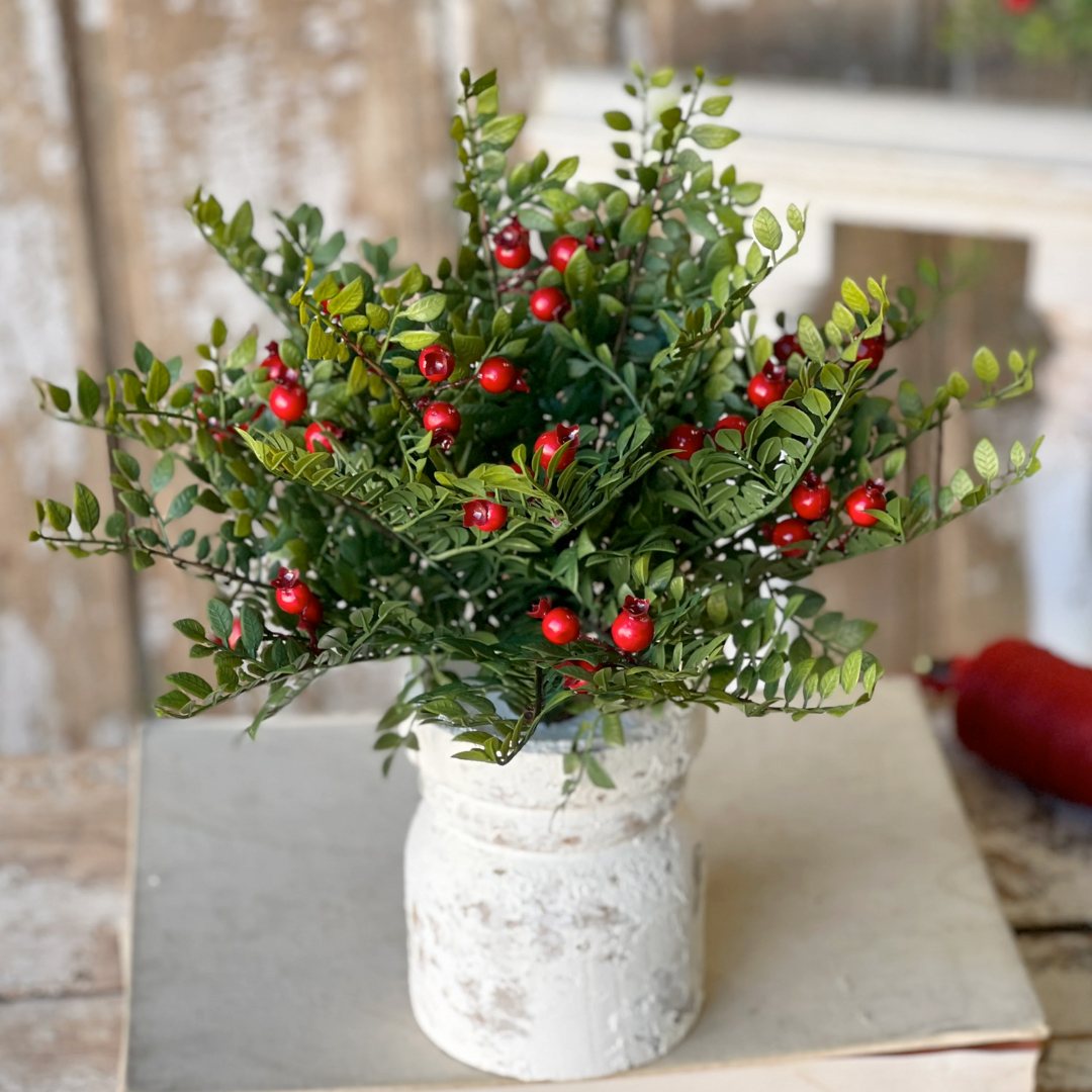 Bouquet of green leaves with red berries in a white vase on a wooden surface.