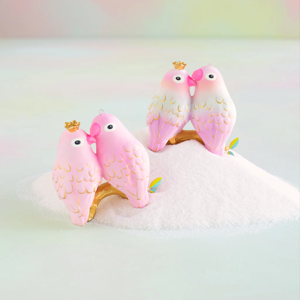Hand-painted resin ornaments in the shape of love birds with pink and white coloring and gold accents, perched on a golden branch, displayed on a surface with white sugar.