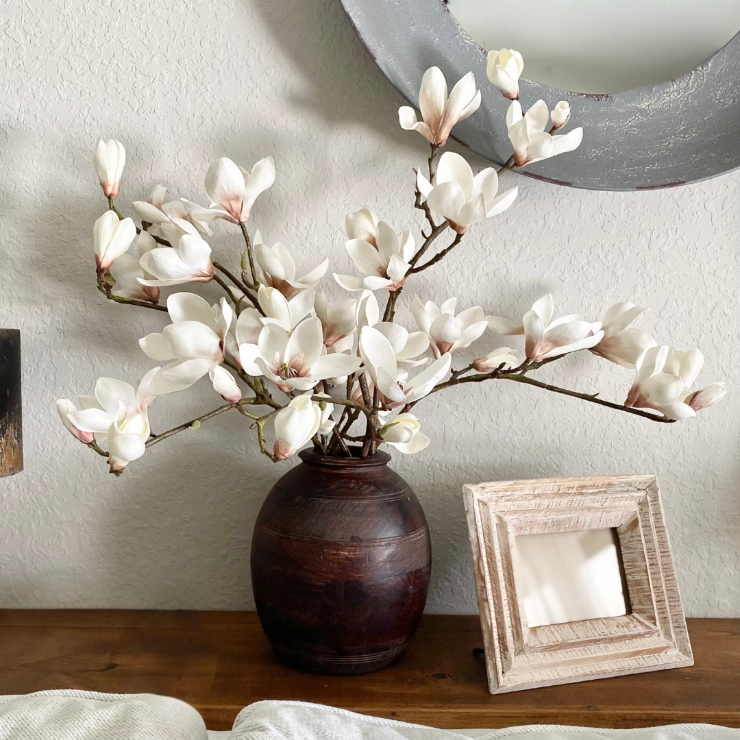 Decorative white magnolia blossoms in a dark vase on a wooden surface with a framed photo in the background.
